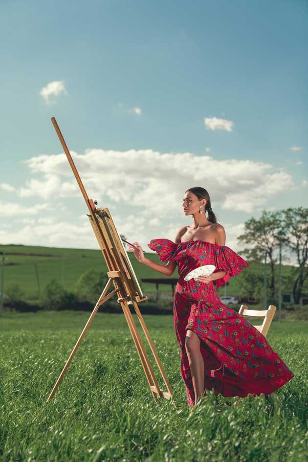 An artistic Eastern European women in elegant red dress painting on a canvas in the field of nature and clear skies